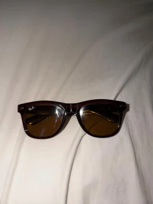 Bruna Wayfarer solglasögon Ray-Ban - Snygga Wayfarer solglasögon från Ray-Ban med bruna bågar och bruna glas. Klassisk och tidlös design som passar till alla outfits. Bågarna är i plast och har Ray-Ban-loggan på sidan.