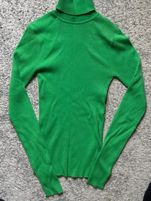 Grön ribbad polotröja - Snygg grön ribbstickad polotröja med lång ärm och figurnära passform. Tröjan har hög krage och är tillverkad i ett stretchigt material som sitter skönt på kroppen. Perfekt för lager på lager och enkel att styla.