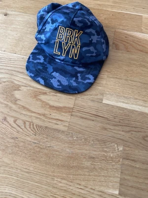 Blå camokeps med BRKLYN-tryck - Säljer en blå keps med camomönster och gul brodyrtext 'BRKLYN' framtill. Kepsen har böjd skärm och justerbar snapback baktill för perfekt passform. Materialet känns lätt och bekvämt, perfekt för en streetwear-look.