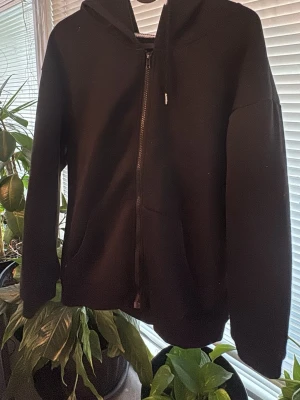 Svart hoodie med dragkedja och huva - Säljer en svart hoodie med dragkedja framtill och stor huva. Hoodien har två fickor på sidorna och är gjord i mjukt, fleeceliknande material på insidan. Perfekt för dig som gillar en enkel och stilren look.
