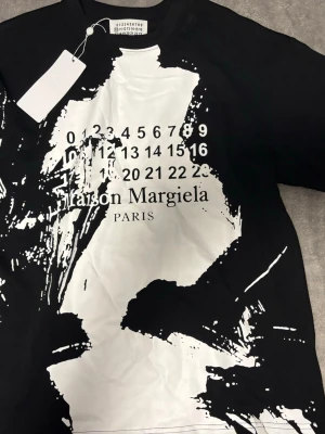 Maison Margiela svartvit t-shirt - Cool svart t-shirt från Maison Margiela med stor vit abstrakt print och ikonisk sifferlogga på bröstet. Oversized fit och rund hals. Tillverkad i Italien, materialet känns mjukt och av hög kvalitet. Perfekt för dig som gillar statement-plagg.