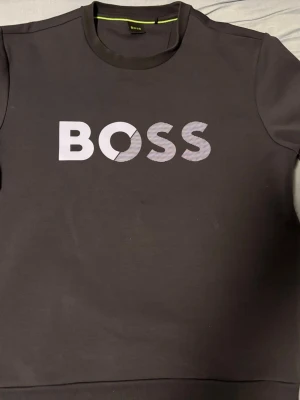 Svart BOSS t-shirt med logga - Snygg svart t-shirt från BOSS med stor vit logga på bröstet. Klassisk rund halsringning och korta ärmar. Perfekt för dig som gillar stilrena och trendiga plagg med tydlig branding.