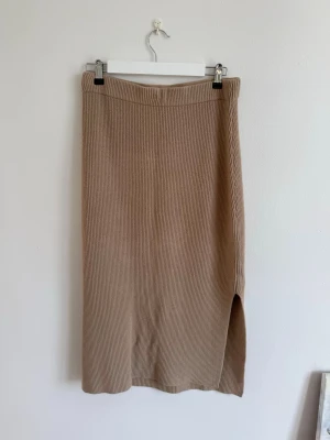 Beige ribbad långkjol med slits - Stilren beige långkjol i ribbat material med en snygg slits på sidan. Kjolen har en rak passform och elastisk midja som gör den bekväm och enkel att bära. 