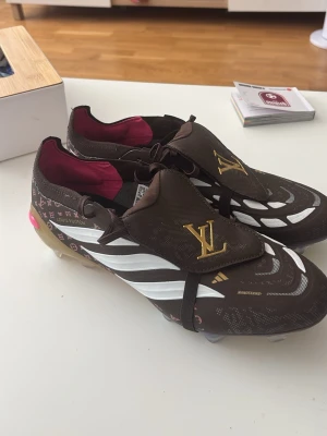 Bruna Louis Vuitton x Adidas fotbollsskor - Unika fotbollsskor i brunt med vita och guldiga detaljer från Louis Vuitton och Adidas. Skorna har klassiska LV-monogram, vita ränder på sidorna och guldfärgad LV-logga på plösen. Insidan är vinröd och yttermaterialet är syntetiskt. Perfekt för dig som vill sticka ut på planen.