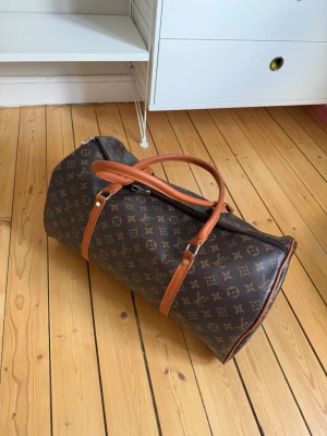 Brun monogram resväska - Snygg brun resväska i klassiskt monogrammönster med ljusbruna läderhandtag och detaljer. Väskan har cylinderform och dragkedja upptill, perfekt för weekendresor.. Materialet är tåligt och lätt att torka av.