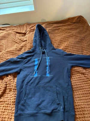 Mörkblå hoodie med blått tryck - Snygg mörkblå hoodie med stor huva och dragsko. Framtill finns ett stort blått tryck i form av två vertikala linjer och en magficka. Hoodien har ribbade muddar vid ärmslut och nederkant. Perfekt för en avslappnad och chill stil.