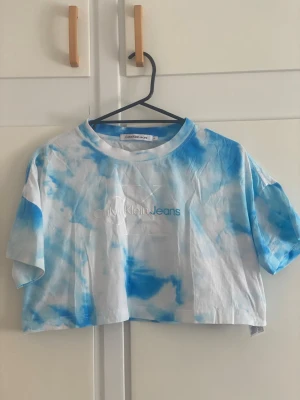 Blåvit cropped t-shirt Calvin Klein - Säljer en blåvit tie-dye cropped t-shirt från Calvin Klein Jeans. T-shirten har rund halsringning och tryckt logga framtill i vitt och blått. Perfekt för en avslappnad och trendig look. Materialet känns mjukt och skönt mot huden.