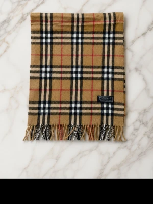  halsduk från Burberry - Klassisk beige halsduk från Burberry med ikoniskt rutmönster i svart, vitt och rött. Halsduken har fransar i båda ändar och är tillverkad i mjukt material som känns lyxigt och stilrent. Perfekt accessoar för att lyfta din outfit.