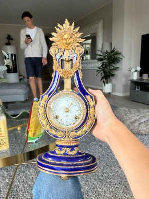 Blå och guldig dekorativ bordsklocka - Säljer en unik bordsklocka i blått och guld med lyraform och detaljerad sol- och ansiktsdekor upptill. Klockan har klassiska romerska siffror och är tillverkad i metall med emaljerad yta. Perfekt som prydnad eller samlarobjekt.