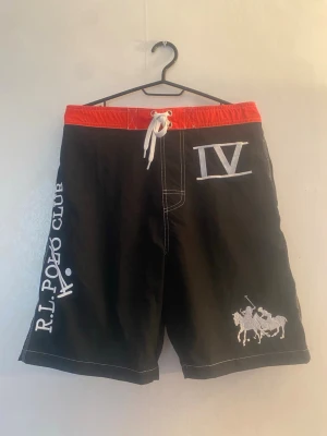 Svarta badbyxor R.L. Polo Club - Snygga svarta badbyxor från R.L. Polo Club med röd midjeresår och vit dragsko. Broderad logga med polospelare, stor IV-detalj och text på benet. Bakficka med knapp och vit kontrastsöm. Perfekta för stranden eller poolen.