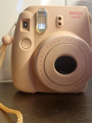 Instax Mini 8 rosa polaroidkamera - Säljer en rosa Instax Mini 8 polaroidkamera med rundad design och inbyggd blixt. Kameran har ett 60mm objektiv och enkel knapp för att ta bilder. Praktisk handledsrem i matchande färg ingår. Perfekt för att ta direktbilder och fånga ögonblick på papper. Lite kantstött men inget som syns allt för mycket