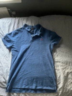Blå pikétröja från Polo Ralph Lauren - Säljer en klassisk blå pikétröja från Polo Ralph Lauren med korta ärmar och krage. Tröjan har knappar vid halsen och den ikoniska röda logotypen broderad på bröstet. Perfekt till jeans eller shorts för en avslappnad look.