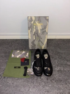 Svarta sneakers med camomönster Valentino - Snygga svarta sneakers med grå och beige camomönster, svarta skosnören och detaljer i mocka och textil. Skorna har en låg siluett och coola detaljer på hälen. Perfekta för dig som vill sticka ut med din stil.