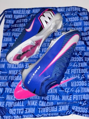 Nike Air Zoom Mercurial fotbollsskor - Säljer ett par Nike Air Zoom Mercurial fotbollsskor i blått med rosa och vita detaljer. Skorna har en strömlinjeformad design, syntetiskt material och dobbar för optimalt grepp på planen. Swoosh-logga på sidan och Mercurial-märke på hälen. Perfekta för snabba spelare.