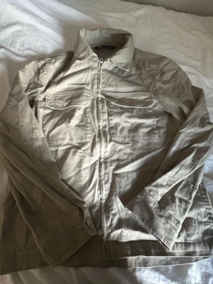 Beige overshirt med dragkedja - Stilren beige overshirt med dragkedja framtill och två stora bröstfickor. Overshirten har klassisk krage och är tillverkad i ett mjukt bomullstyg. Perfekt att slänga över en t-shirt för en avslappnad look.