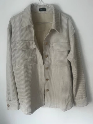 Beige overshirt i manchester från Lindex - Stilren beige overshirt från Lindex i mjuk manchester. Skjortan har klassisk krage, bröstfickor med lock och knappar framtill samt vid ärmslut
