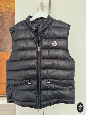 Svart dunväst från Moncler - Snygg svart dunväst från Moncler med klassisk quiltad design och hög krage. Västen har två dragkedjefickor framtill och Monclers ikoniska logga på bröstet. Perfekt för lager-på-lager och håller dig varm med stil.