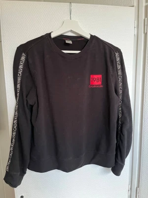 Svart loungewear-tröja Calvin Klein - Svart loungewear-tröja från Calvin Klein med tryckt logga och texten '1981' i rött på bröstet. Långa ärmar med Calvin Klein 1981-tryck längs båda ärmarna. Rund halsringning och mjukt material som passar perfekt för chill dagar hemma.