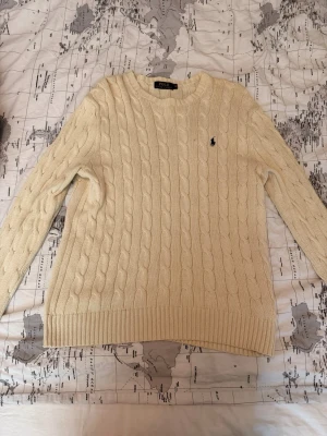 Kabelstickad offwhite tröja Polo Ralph Lauren - Kabelstickad offwhite tröja från Polo Ralph Lauren med rund halsringning och den klassiska broderade loggan på bröstet. Tröjan har långa ärmar och ribbade muddar vid ärmslut och nederkant. Perfekt för lager på lager och en clean look.