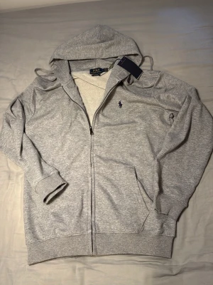 Grå hoodie från Polo Ralph Lauren - Snygg grå hoodie från Polo Ralph Lauren med dragkedja framtill och klassisk blå logga broderad på bröstet. Tröjan har huva med dragsko, ribbade muddar och fickor på sidorna. Tillverkad i mjuk bomullsblandning för skön komfort.