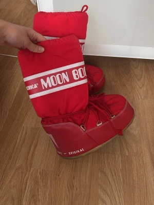 Röda Moon Boot Original boots - Säljer ett par ikoniska röda Moon Boot Original boots med vit logga och snörning framtill. De har en klassisk rund form, högt skaft och är tillverkade i syntetmaterial med mjuk insida. Perfekta för vinterns alla äventyr och håller fötterna varma. Skulle säga att dem passar en 38-40 i storlek då den anpassar sig bra efter foten.