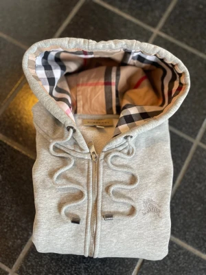 Grå zip hoodie från Burberry - Snygg grå hoodie med dragkedja från Burberry. Klassisk huva med det ikoniska rutmönstret på insidan, dragsko och två fickor framtill. Tillverkad i mjukt bomullsmaterial och har diskret Burberry-logga broderad på bröstet. Perfekt för en avslappnad men stilren look. Dragkedjan är sönder men det går lätt att fixa hemma eller hos en sömmerska annars mycket bra skick 