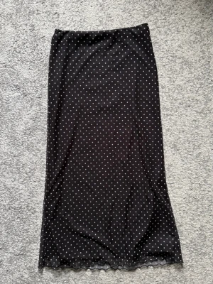 Svart prickig långkjol - Snygg svart långkjol med vita prickar. Kjolen har en rak och enkel siluett som ger en stilren look. Perfekt att matcha med en enkel topp eller stickad tröja. Materialet känns lätt och följsamt.