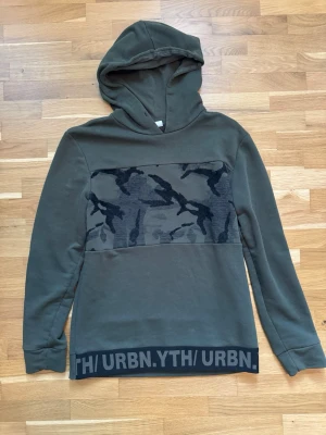 Jack & Jones Olivgrön hoodie med camomönster - Snygg olivgrön hoodie med huva och lång ärm. Framtill finns ett brett camomönstrat parti i svart och grått samt en svart kant längst ner med texten 'URBN.YTH'. Perfekt för dig som gillar streetwear och vill ha en avslappnad stil.