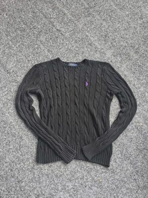 Svart kabelstickad tröja Polo Ralph Lauren - Riktig kabelstickad svart tröja från Polo Ralph Lauren med lila broderad logga på bröstet. Tröjan har rund halsringning, långa ärmar och ribbade muddar. Klassisk och tidlös design som passar till mycket.