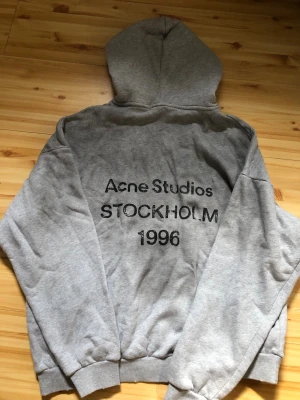 Grå hoodie Acne Studios Stockholm - Snygg grå hoodie från Acne Studios med trycket 'Acne Studios STOCKHOLM 1996' på ryggen. Klassisk passform med huva och ribbade muddar. Perfekt för dig som gillar streetwear och vill ha något stilrent och bekvämt. Materialet är mjuk bomullsblandning.