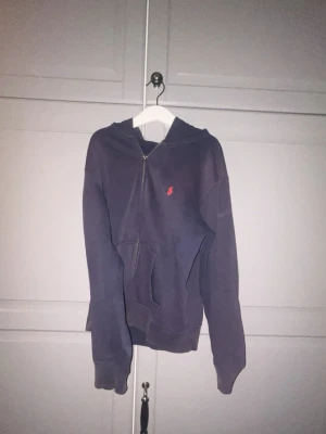 Mörkblå hoodie med dragkedja - Snygg mörkblå hoodie med dragkedja och huva. Har en röd liten logga broderad på bröstet och praktisk känguruficka framtill. Perfekt för en avslappnad och cool stil.