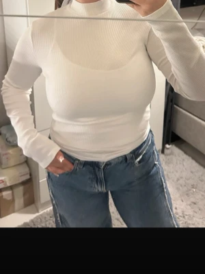 Vit ribbad topp med öppen rygg och knyt - Säljer en vit långärmad ribbad topp med hög hals. Toppen har en snygg öppen rygg med knytband både i nacken och nedtill, vilket ger en unik och trendig look. Perfekt att styla med jeans eller kjol för en cool outfit.