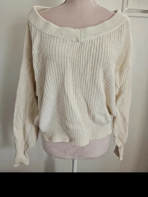 Off shoulder stickad tröja cremevit - Trendig off shoulder stickad tröja i cremevitt med bred ribbad kant vid halsen. Tröjan har en lös och avslappnad passform, långa ärmar och ribbade muddar. Perfekt för lager på lager och chill stil.