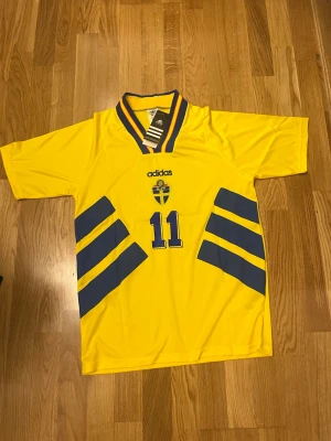Svensk landslagströja Brolin #11 Adidas - Gul svensk landslagströja från Adidas med blå detaljer, nummer 11 och namnet Brolin på ryggen. Klassisk v-ringad krage med blå och gula ränder, samt det svenska landslagsemblemet på bröstet. Perfekt för fotbollsfans!