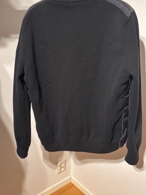 Svart cardiganjacka från Moncler - Snygg svart cardiganjacka från Moncler med stickade ärmar och rygg samt vadderad framsida. Jackan har dragkedja, hög krage och Moncler-logga på ärmen. Perfekt för lager-på-lager och en stilren look.
