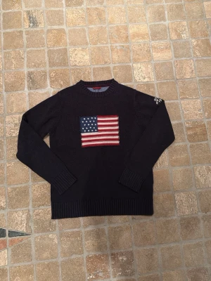Mörkblå stickad tröja med USA-flagga - Snygg mörkblå stickad tröja med en stor amerikansk flagga på bröstet. Tröjan har rund halsringning, ribbade muddar och detaljer med text på ena ärmen. Perfekt för dig som gillar klassisk och avslappnad stil.