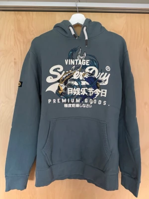 Superdry hoodie - Snygg hoodie från Superdry med stort grafiskt tryck och japansk text på bröstet. Klassisk känguruficka framtill, ribbade muddar och vit dragsko i huvan. Perfekt för en avslappnad streetwear-look.