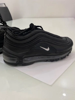 Nike Air Max 97 svarta sneakers - Säljer ett par svarta Nike Air Max 97 sneakers med ikoniska vågiga paneler och synlig Air-enhet i sulan. Väldigt sparsamt använda! I storlek 35.5, som någon som bär skostorlek 36/35 har jag kunnat bruka dessa. Självklart äkta! 