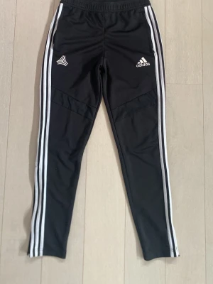 Svarta Adidas träningsbyxor herr - Säljer ett par svarta träningsbyxor från Adidas med klassiska vita ränder längs benen och logga på vänster sida. Byxorna har smal passform, resår i midjan och är tillverkade i ett lätt och stretchigt material som passar perfekt till träning.