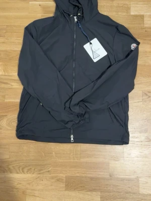 Svart vindjacka med huva Moncler - Snygg svart vindjacka från Moncler med huva och dragkedja framtill. Jackan har två fickor med tryckknappar, elastiska muddar och Moncler-logga på ärmen. Huvan har en rödvit rand som detalj. Perfekt för blåsiga dagar.