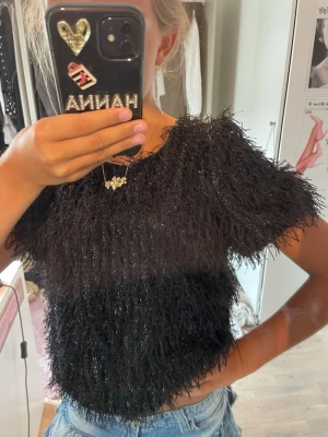 Svart glittrig topp med fluffig textur - Säljer en svart glittrig topp med korta ärmar och fluffig . Toppen har en croppad passform och ger en cool statement-look. Perfekt för dig som vill sticka ut och älskar trendiga detaljer.