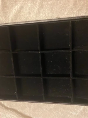 Svart smyckesförvaring med fack - Praktisk svart smyckesförvaring med flera fyrkantiga fack, perfekt för att organisera ringar, örhängen , klockor eller andra små accessoarer. Tillverkad i ett mjukt material som skyddar dina saker och håller dem på plats.