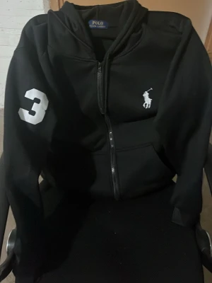 Svart hoodie från Polo Ralph Lauren - Svart hoodie med dragkedja från Polo Ralph Lauren. Stort vitt nummer 3 på ena ärmen och klassisk broderad logga på bröstet. Huva och fickor framtill, perfekt för en avslappnad och cool look.