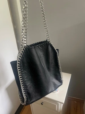 Svart handväska med kedjedetaljer - Snygg svart handväska i fejkläder med grova silverfärgade kedjor som axelband och längs kanterna. Väskan har en mjuk, lite slouchy form och passar perfekt till en trendig vardagslook. Enkel att matcha med olika outfits och rymmer det viktigaste.