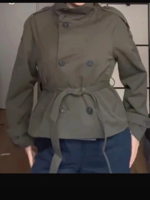 Olivgrön kort trenchcoat med bälte - Snygg olivgrön trenchcoat med dubbelknäppning och knytskärp i midjan. Jackan har axelklaffar och hög krage, perfekt för en trendig look. Klassisk design med moderna detaljer.
