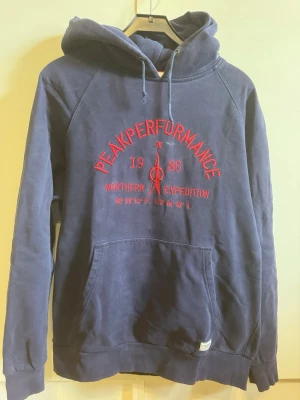 Mörkblå Peak Performance hoodie - Snygg mörkblå hoodie från Peak Performance med röd brodyr framtill där det står 'Peak Performance 1986 Northern Expedition'. Klassisk känguruficka och justerbar huva med dragsko. Perfekt för en avslappnad och sportig stil.