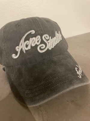 Grå keps från Acne Studios - Snygg grå keps från Acne Studios med broderad logga i vitt framtill och på skärmen. Klassisk böjd skärm och justerbar rem bak. Tillverkad i mjukt bomullstyg som ger en avslappnad och trendig look.