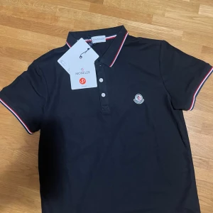 Svart Moncler pikétröja med röd/vit kant - Klassisk svart pikétröja från Moncler med korta ärmar och knappar framtill. Tröjan har röd och vit rand längs krage och ärmslut samt Moncler-logga på bröstet. Tillverkad i mjuk bomull för en bekväm känsla.
