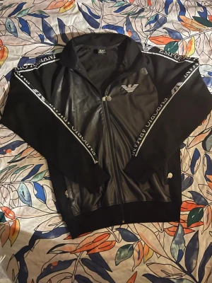 Svart hoodie från Emporio Armani - Snygg svart hoodie från Emporio Armani med dragkedja framtill och logga på bröstet. Har vita detaljer och text längs ärmarna samt ribbade muddar. Perfekt för en stilren och sportig look.