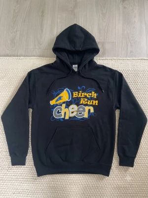 Svart Gildan hoodie Birch Run Cheer - Svart hoodie från Gildan med trycket 'Birch Run Cheer' i gult, blått och vitt framtill samt en megafon-grafik. Klassisk känguruficka och dragsko i huvan. Tillverkad i tjockt material, perfekt för dig som gillar cheerleading och streetwear.